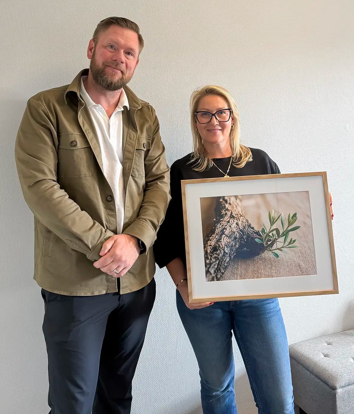 Tobias Petersson och Helen Christensen står bredvid varandra mot en ljus vägg och ler mot kameran. Tobias till vänster i olivgrön skjortjacka över vit skjorta, Helen till höger med blont axellångt hår, mörka glasögon, svart tröja och jeans. Hon håller en inramad bild av en grov trädgren med en liten grön kvist.