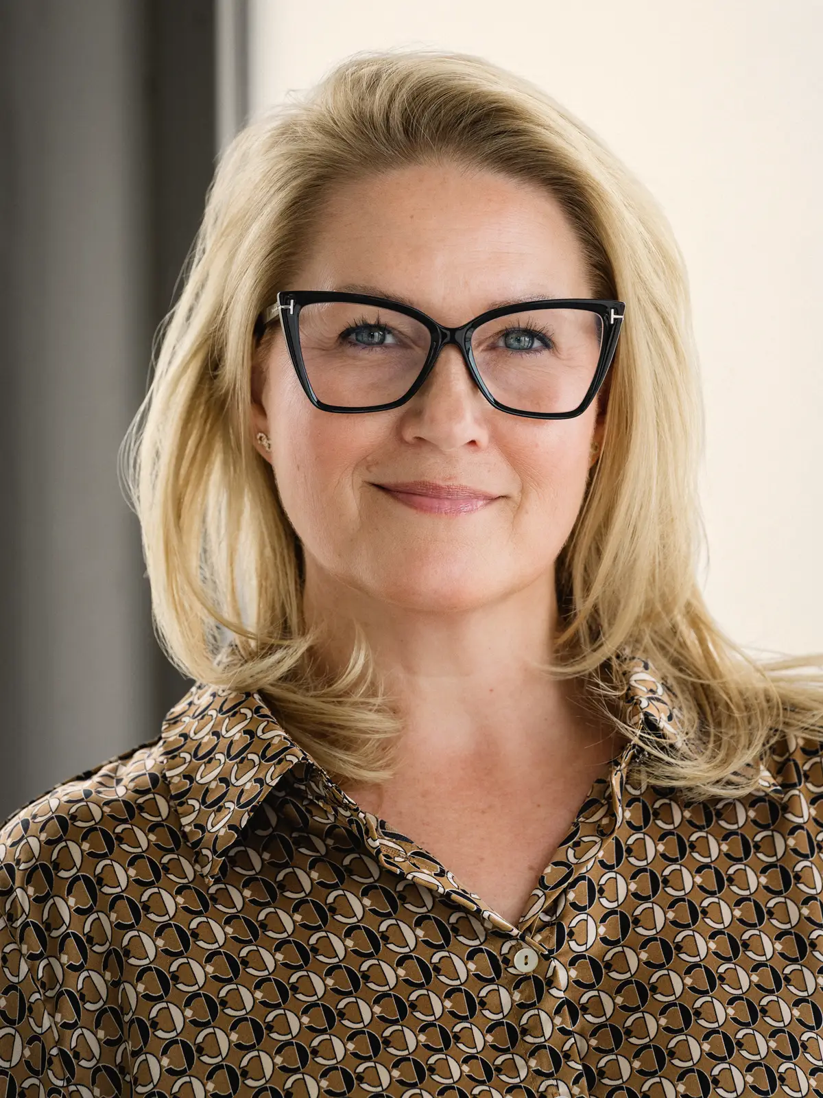 Helen Christensen fotograferad inomhus mot en ljus vägg. Halvbild med blont axellångt hår, mörka glasögon med kantiga bågar och en mönstrad skjorta i brunt, svart och beige. Hon ser in i kameran med ett lätt leende.